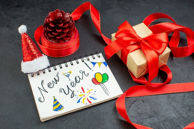 Free photo top view of a gift conifer cone with red ribbon and notebook with new year writing and santa claus hat beautiful gift on dark background