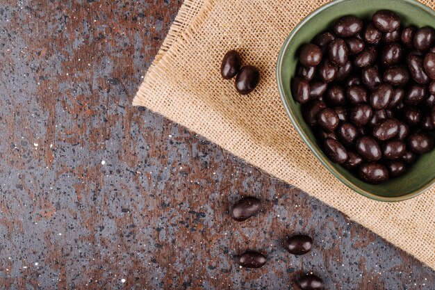 Top view of glazed chocolate nut candy in a bowl on sackcloth on rustic background with copy space