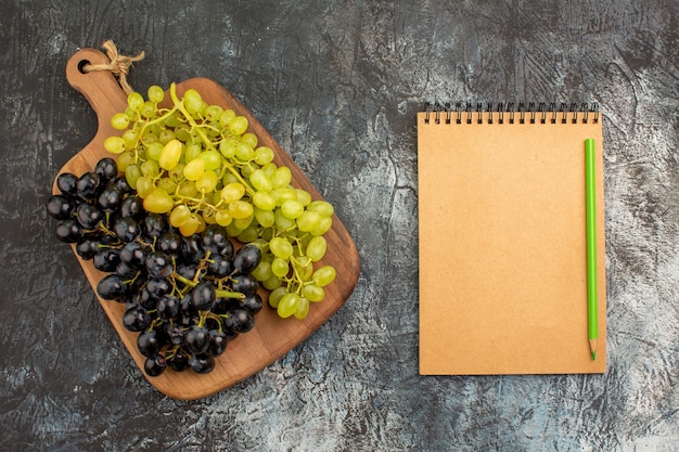 Free photo top view grapes notebook pencil bunches of green and black grapes on the cutting board