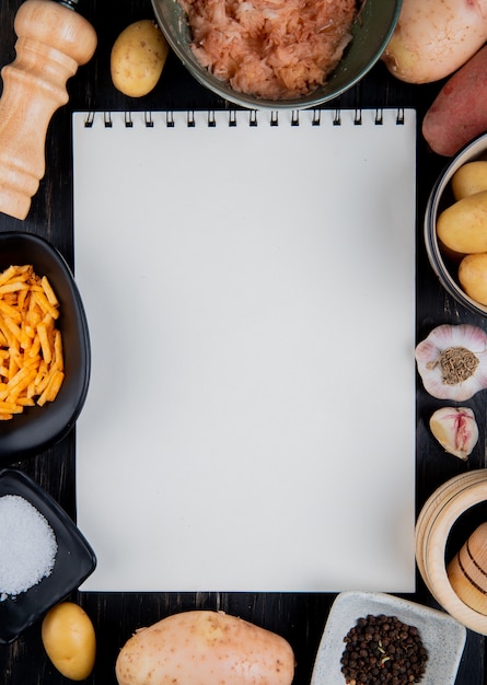 Top view of grated sliced and whole potatoes around note pad with salt black pepper on wooden surface with copy space