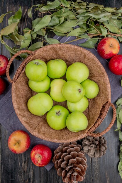 Top view of green apples in basket with red apples pinecones and leaves on cloth and wooden table