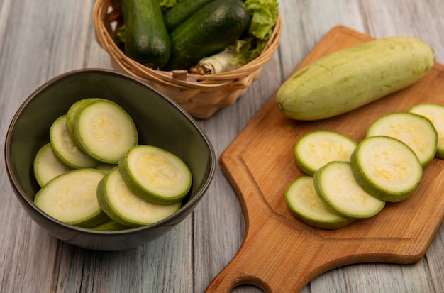 Top view of green skinned zucchinis on a wooden kitchen board with chopped zucchinis on a bowl with cucumbers and lettuce on a bucket on a grey wooden wall