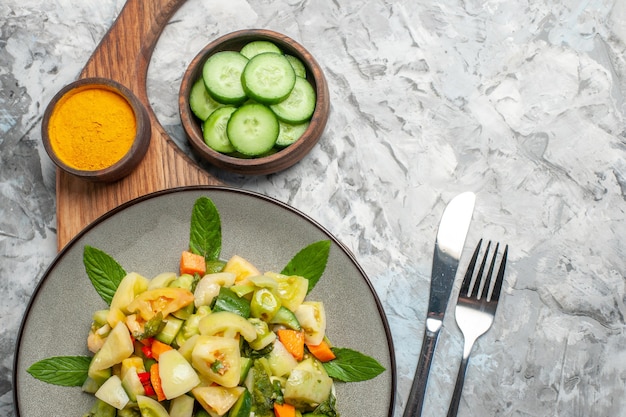 Top view green tomato salad on oval plate turmeric on cutting board bowl with cut cucumber fork and knife on dark background