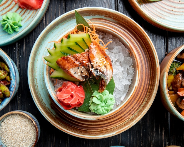 Top view of grilled japanese eel with sliced cucumbers and gingers served with wasabi sauce on bamboo leaf and on ice cubes in a plate on the table