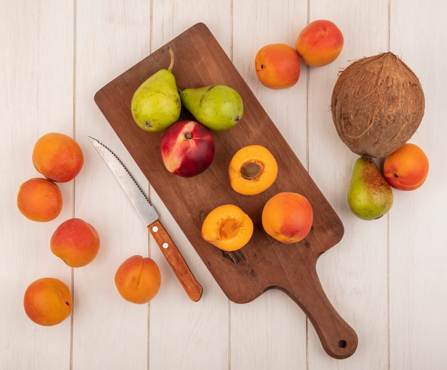 Top view of half cut and whole fruits as peach apricots and pears on cutting board and pattern o apricots pear and coconut and knife on wooden background