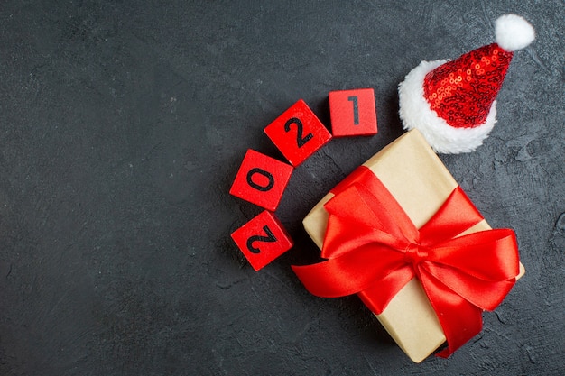 Top view of hand holding beautiful gift with bow-shaped ribbon next to santa claus hat numbers on dark background