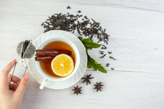 Top view of hand holding black tea in a white cup around dry tea and leaves on white table