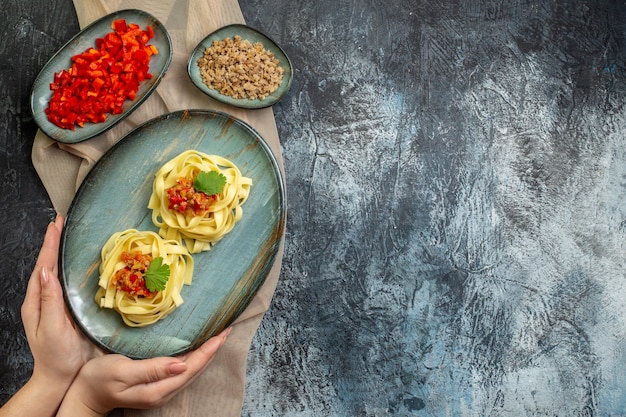 Free photo top view of hand holding a blue plate with delicious pasta meal served with tomato and meat for dinner on tan color towel its ingredients