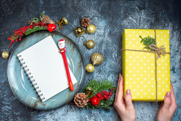 Top view of hand holding a gift box and notebook with pen blue plate decorative accessories conifer cone on dark background