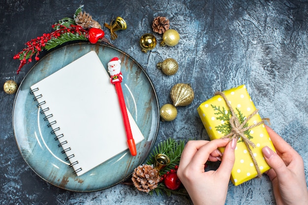 Top view of hand opening a gift box and notebook with pen blue plate decorative accessories conifer cone on dark background