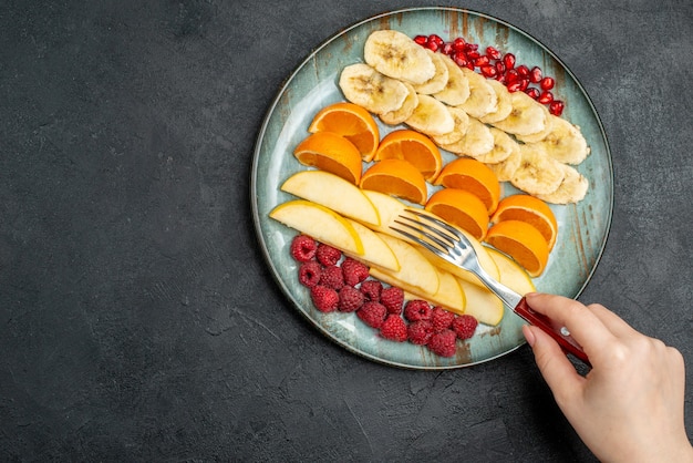 Top view of hand taking apple slices from collection of chopped fresh fruits on a blue plate on black table