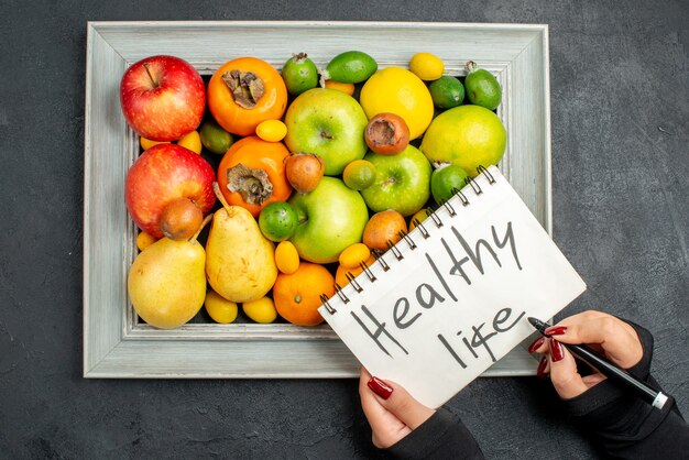 Top view of hand writing healthy life on spiral notebook on collection of fresh fruits in picture frame on dark background