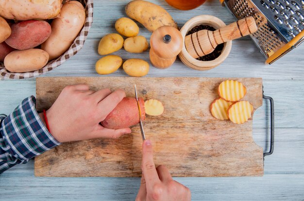 Top view of hands cutting potato with knife on cutting board with other ones in basket with black pepper seeds grater and other potatoes on wood