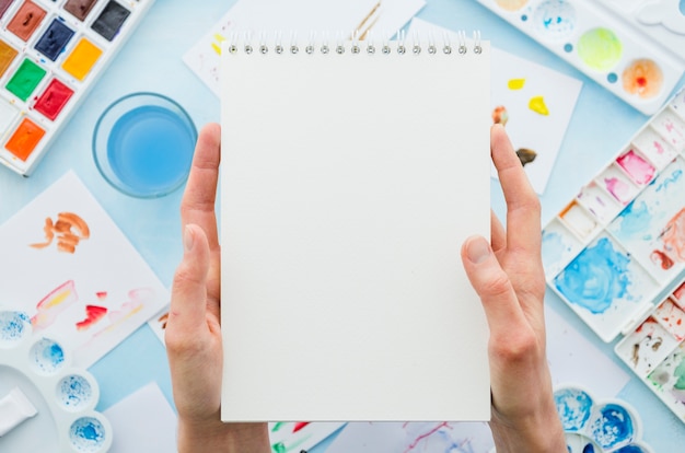 Top view hands holding notebook surrounded by painting elements