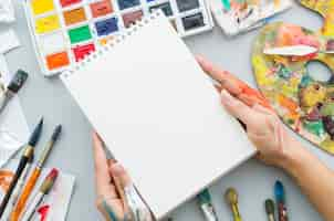 Free photo top view hands holding notebook surrounded by painting elements