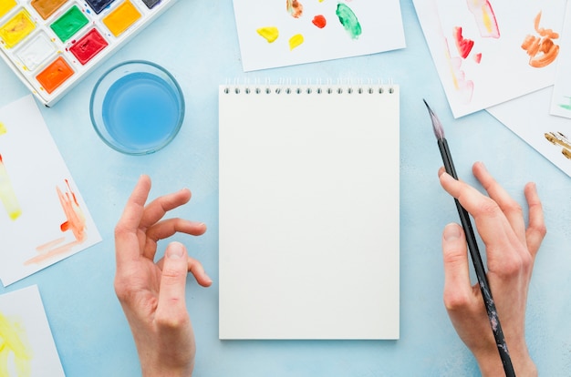 Free photo top view hands and notebook surrounded by painting elements