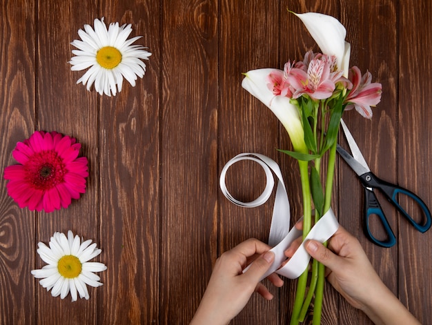 Free photo top view of hands tying with a ribbon a bouquet of pink and white color alstroemeria and calla lilies flowers on wooden background