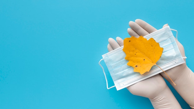 Free Photo top view of hands with gloves holding medical mask with autumn leaf