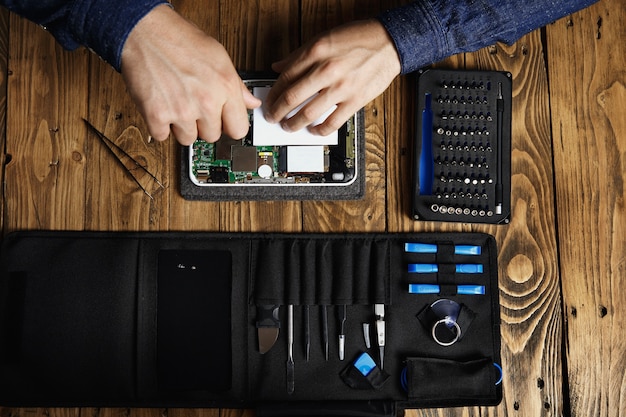 Top view of hands works on broken electronic gadget to fix it near tool bag and on wooden table in service shop