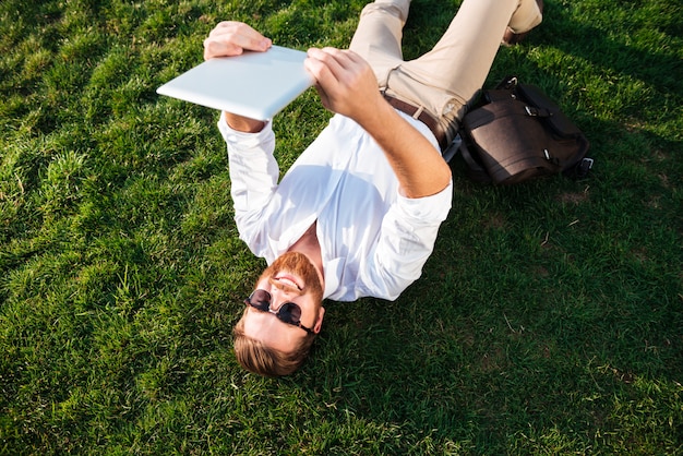 Free photo top view of happy bearded man in sunglasses and business clothes lying on grass outdoors and making selfie on tablet computer