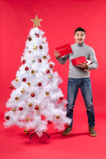 Top view of happy handsome adult in a gray blouse standing near the decorated white Christmas tree