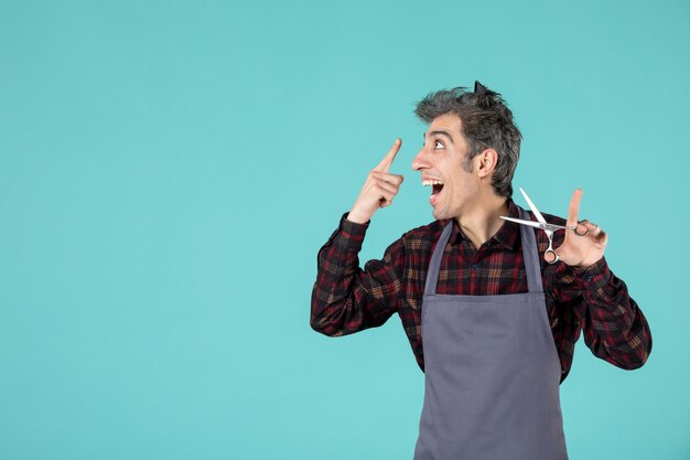 Top view of happy satisfied young barber wearing gray apron and holding comb on his head and showing scissor pointing something on the right side on blue color background