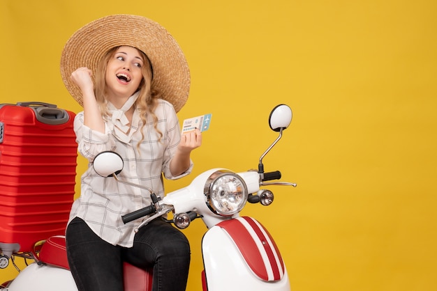 Top view of happy young woman wearing hat and sitting on motorcycle and holding ticket on yellow 