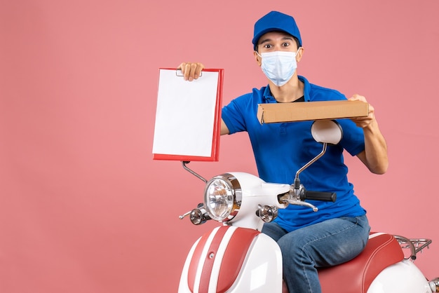 Free Photo top view of hardworking courier guy in medical mask wearing hat sitting on scooter holding document on pastel peach background