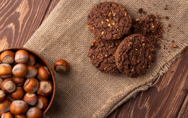 Top view of hazelnuts in a wooden bowl and oatmeal cookies on sackcloth on wooden