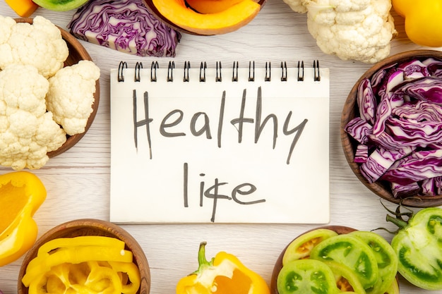 Free Photo top view healthy life written on notebook red cabbage cauliflower yellow bell pepper green tomato in bowls on white wooden surface