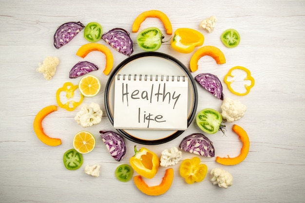 Free photo top view healthy life written on notepad on round platter cut vegetables on white wooden surface