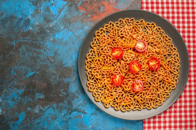 Top view heart shaped pasta with cherry tomatoes on dark oval platter on blue table copy place