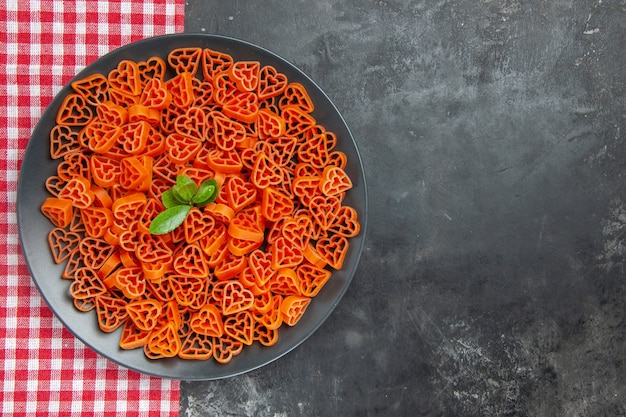 Free photo top view heart shaped red italian pasta on black oval plate on kitchen towel on dark table with free space