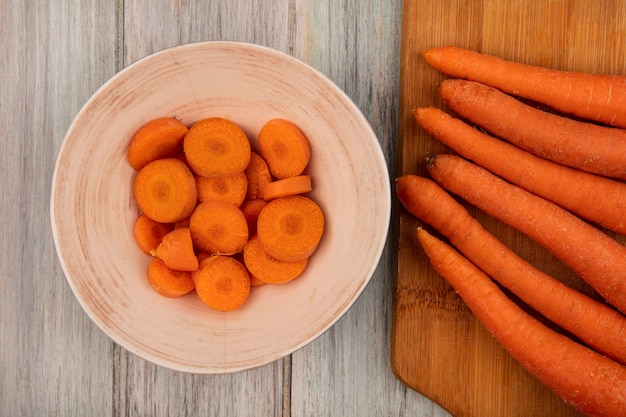 Top view of highly nutritious carrots on a wooden kitchen board with chopped carrots on a bowl on a grey wooden wall