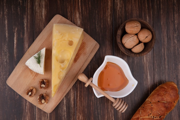 Top view honey in a saucer with a variety of cheeses on a stand with walnuts and a loaf of bread on a wooden background