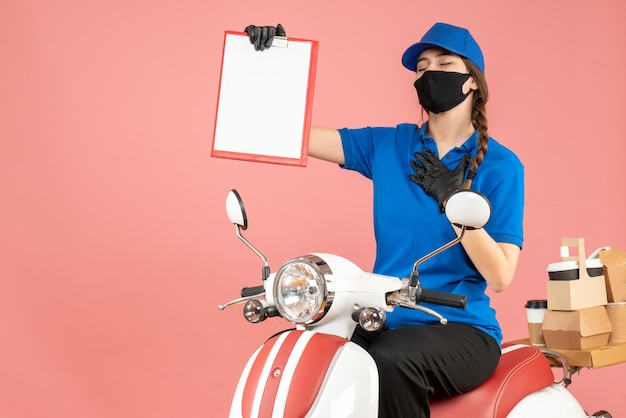 Free photo top view of hopeful courier girl wearing medical mask and gloves sitting on scooter holding empty paper sheet delivering orders on pastel peach background