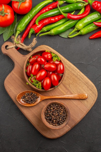 Free photo top view hot red and green peppers and tomatoes bay leaves bowls with cherry tomatoes and black pepper and spoon on a chopping board on black ground