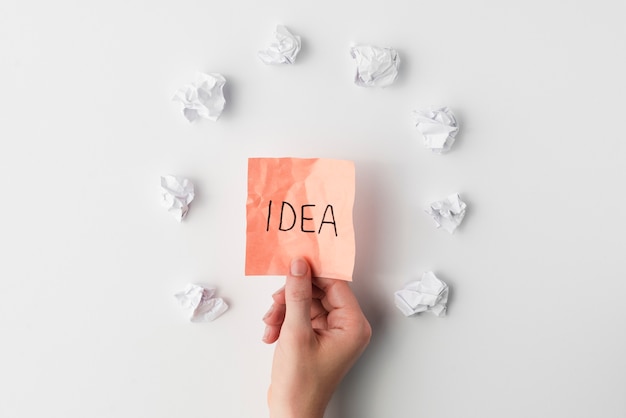 Top view of human hand holding sticky note with idea text surrounded by crumpled paper on white background