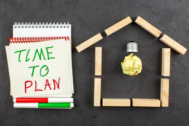 Top view idealight bulb in house shaped wood blocks time to plan written on notepad red and green markers on black