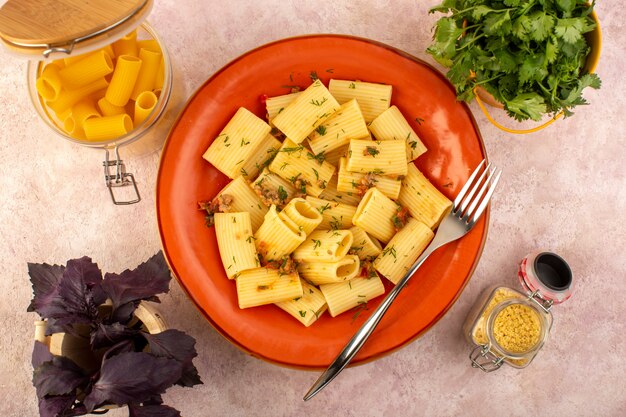 A top view italian pasta cooked tasty salted inside round orange plate with flower and raw pasta on pink desk