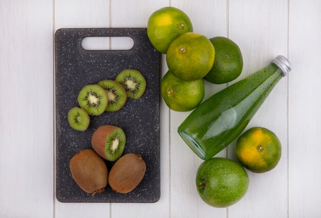 Top view kiwi on cutting board with tangerines apple and bottle of juice on white wall