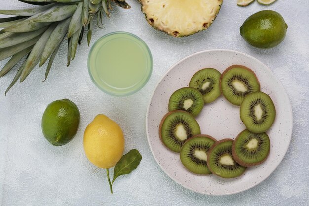 Top view of kiwi slices in plate with lime juice in glass pineapple slices lime and lemon and pineapple leaves on white background