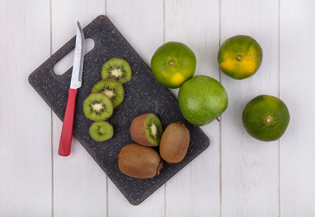 Top view kiwi with knife on cutting board with tangerines and apple on white wall