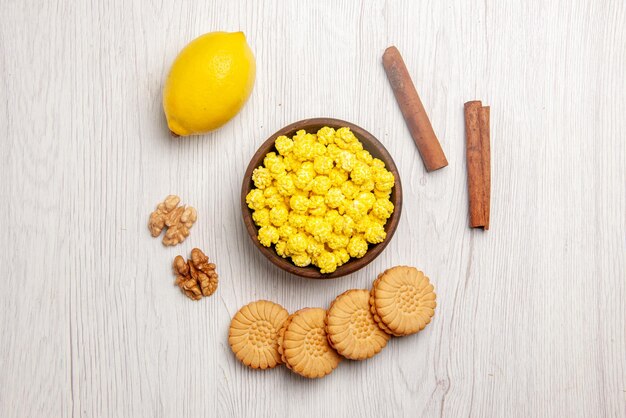 Top view lemon and sweets cinnamon sticks cookies nuts bowl of yellow candies and lemon on the white table