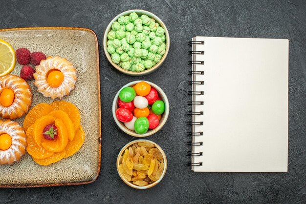 Top view of little cakes with lemon slices tangerines and candies on black table