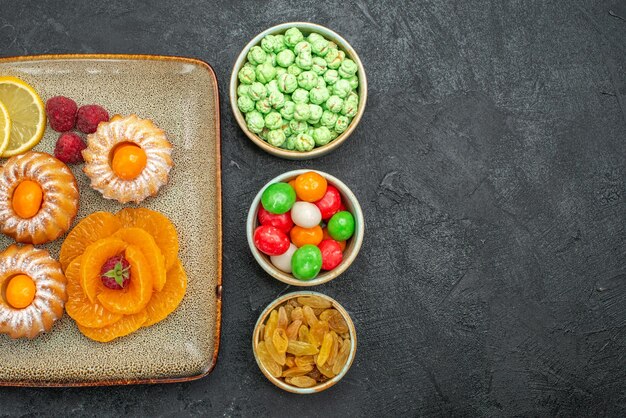 Top view of little cakes with lemon slices tangerines and candies on black