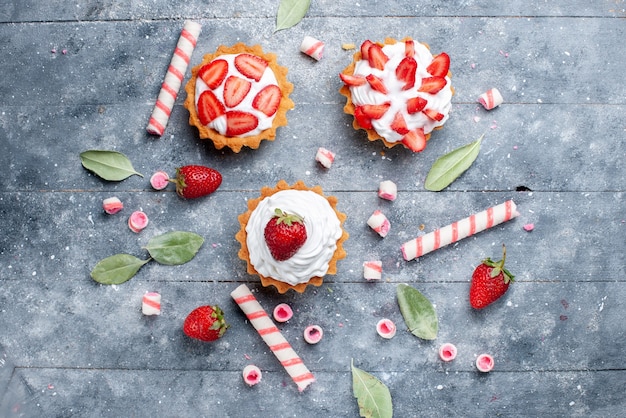 Top view little creamy cakes with sliced and fresh strawberries along with stick candies on the grey surface fruit cake sweet color bake berry