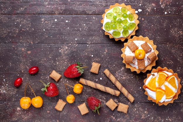 Free Photo top view little creamy cakes with sliced grapes oranges along with strawberries on the brown wooden desk cake biscuit fruit mellow