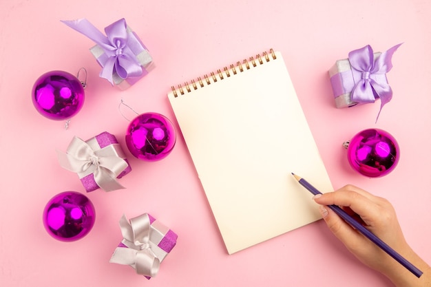 Top view of little presents with xmas tree toys and notepad on pink surface