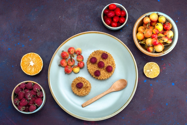 Top view of little round cake with fresh raspberries inside plate with fresh fruits on dark surface